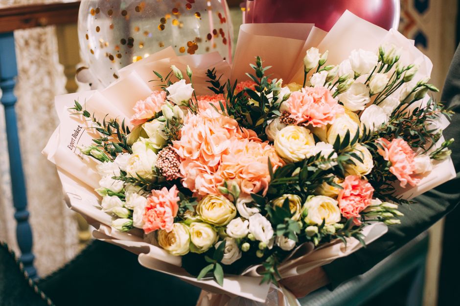 A vibrant bouquet arrangement featuring soft peach and white roses, complemented by delicate white lisianthus and sprigs of green foliage, wrapped in elegant light pink paper with a white ribbon. The
