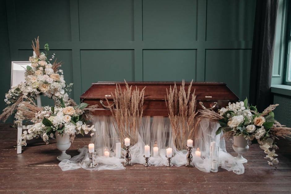 A funeral flower arrangement featuring two large white floral urns filled with cream and white roses, hydrangeas, and greenery, positioned on either side of a vintage wooden casket, all set on a dark 