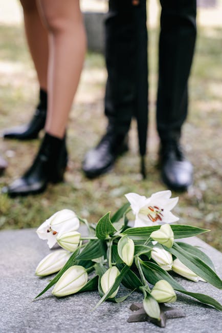 A bouquet of white lilies with open blooms and closed buds, arranged with long green leaves on a flat surface outdoors. In the background, a person in dark trousers and polished black shoes stands on 