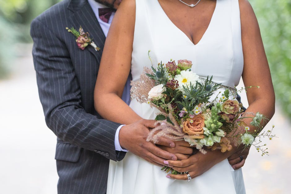 Close-up of a bride holding a floral bouquet featuring peach roses, white daisies, and green foliage, with a groom standing behind her, dressed in a dark striped suit with a pin in his lapel. The bouq