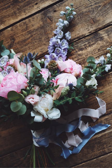 A bouquet of fresh flowers arranged with soft pink peonies, white roses, and sprigs of eucalyptus, complemented by tall delicate blue lavender and white stock flowers, all wrapped in elegant satin rib