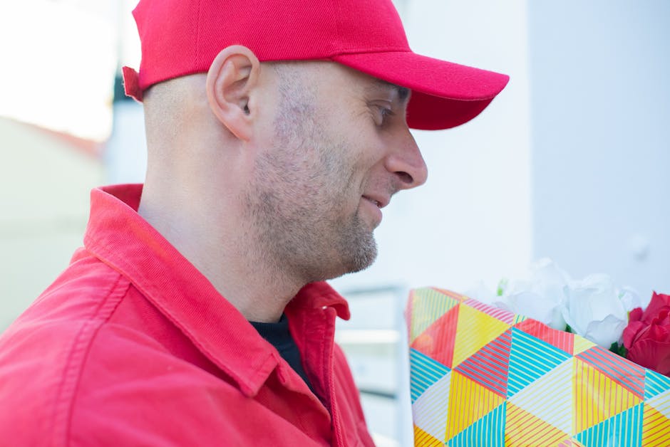 A man wearing a red cap and red jacket holds a vibrant, a geometrically patterned gift bag containing fresh flowers, including white roses and pink blooms, wrapped in white tissue paper. The setting a