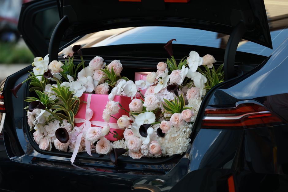 A car's open trunk filled with a lush floral arrangement comprising white and blush pink roses, white orchids, dark burgundy calla lilies, and green-leaved foliage, surrounding a pink gift box tied wi
