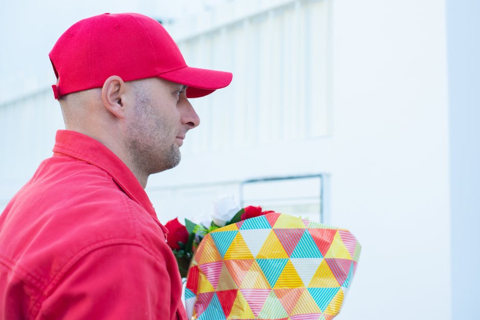 A man wearing a red cap and matching red jacket holds a floral gift wrapped in colorful geometric-patterned paper with yellow, blue, pink, and orange triangles. He is pictured in side profile, with a