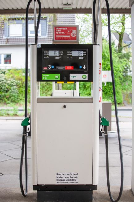 The image shows a fuel pump at a petrol station with a digital display screen indicating fuel prices and options. The pump is white with green and black accents, featuring two black hoses with green h