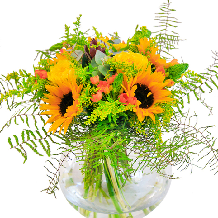 The initial impression of this autumn-inspired bouquet is pure exuberance-a glowing arrangement that channels the golden hours of Leyton life. Large, sunny sunflowers and soft yellow roses rest side by side, their petals layered in shining abundance, casting the suggestion of sunbeams captured in floral form. Between these radiant core blooms, lush green leaves and trailing, airy foliage spill outward, echoing the untamed beauty found in Leyton's plentiful pocket parks. Clusters of deep red Hypericum berries nestle amid the leafage, their glossy skins popping against the soft, matte textures of the surrounding flowers. The entire collection is grounded in a smooth, round glass vase, its clarity revealing the vibrant stems converging below-a subtle tribute to both simplicity and architectural grace. Drawing in closer, one can almost feel the textures: velvety sunflower discs, satiny rose petals, crisp new growth, and the cool touch of eucalyptus. Faint whispers of fragrance drift up-sweetly fresh, hinting at wild fields and local gardens dappled in sunlight. This bouquet is more than decoration; it's a heartfelt gesture suited for birthdays celebrated on Church Road, anniversaries shared above Leyton Midland, or simply brightening an everyday moment within the neighbourhood. Designed thoughtfully by Leyton Florist, it's a centrepiece that floods any room with joy, hope, and the spirit of celebration found on Leyton's brightest days.