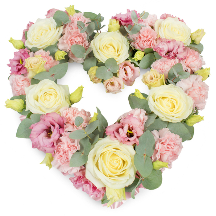 A luxuriously full, heart-shaped funeral tribute appears against a pure white backdrop, its carefully crafted form radiating warmth and tenderness. The heart is entirely covered with flowers, creating a continuous surface of soft colour and texture with no gaps, as though the blossoms themselves are forming a gentle embrace. Dominating the design are large, beautifully formed roses in creamy ivory and pale buttery yellow, their tightly spiralled centres opening into layered, velvety petals that lend the arrangement an air of elegance and purity. Between these striking roses, clusters of light, pastel pink carnations fill the spaces, their ruffled, multi-petalled heads adding a rich, tactile softness, like folded fabric. Deeper tones of rose-pink and mauve lisianthus are scattered throughout, their more open, trumpet-like blooms and occasionally darker throats drawing the eye inward, suggesting the deeper currents of emotion behind the tribute. Cool, silvery-green eucalyptus foliage is woven delicately among the blossoms, its rounded leaves peeking from beneath petals, defining the crisp heart outline and providing a calm, natural contrast to the pastel palette. Small, yellow-green buds appear in places, their youthful tightness hinting at the cycle of life continuing. As a piece often created for families in Leyton and the surrounding streets off Lea Bridge Road, this heart wreath feels deeply personal, suitable for resting near a coffin, on a stand during a memorial service, or by a photograph at a wake. It conveys quiet sympathy, remembrance, and an enduring promise of love that stays long after the ceremony is over.